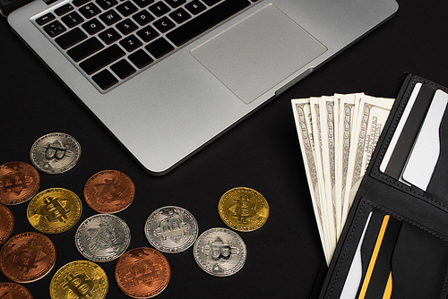 KYIV, UKRAINE - APRIL 26, 2022: Top view of wallet with money near laptop and bitcoins on black background