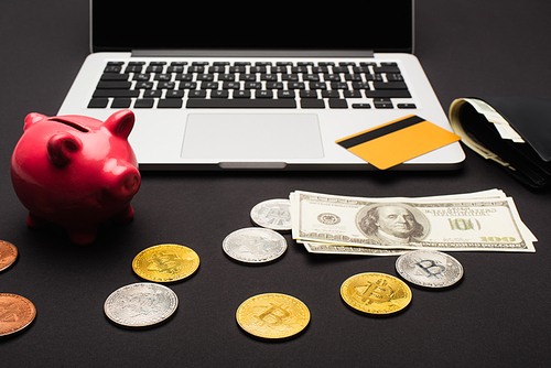 KYIV, UKRAINE - APRIL 26, 2022: Bitcoins near dollars and laptop on black background