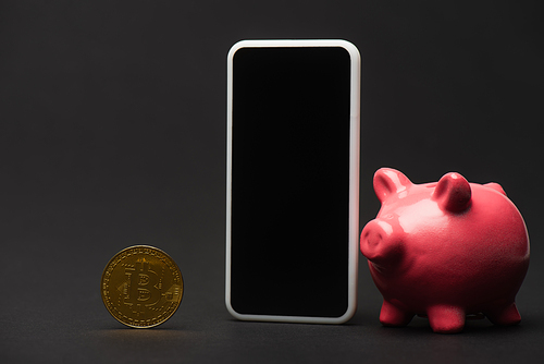 KYIV, UKRAINE - APRIL 26, 2022: Close up view of smartphone near piggy bank and bitcoin on black background