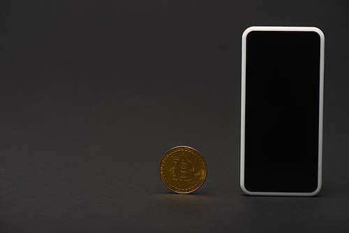 KYIV, UKRAINE - APRIL 26, 2022: Golden bitcoin and cellphone with blank screen on black background