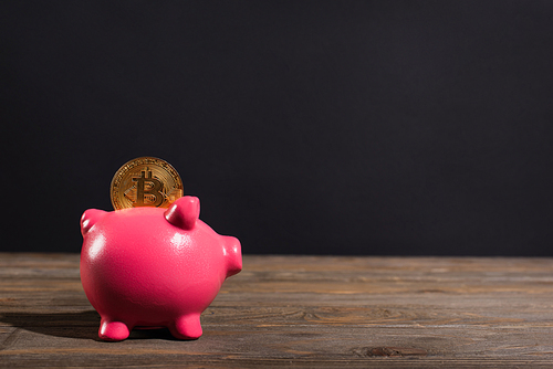 KYIV, UKRAINE - APRIL 26, 2022: Golden bitcoin on piggy bank on wooden surface on black background