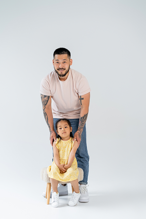 full length of smiling asian man with tattoos standing near preschooler daughter on grey