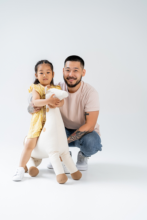 full length of happy asian kid holding toy giraffe and standing near tattooed father on grey