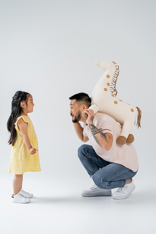 side view of tattooed asian man holding toy giraffe near preschooler daughter on grey