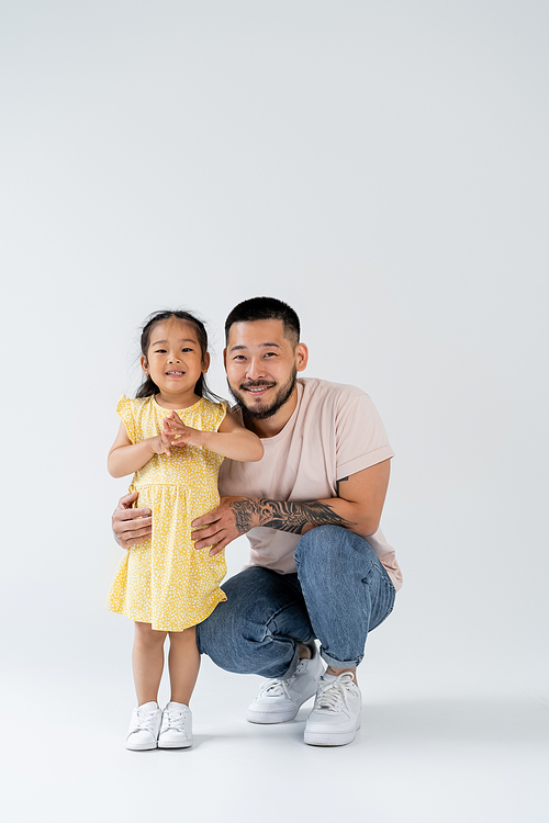 full length of happy and tattooed asian father hugging preschooler daughter on grey