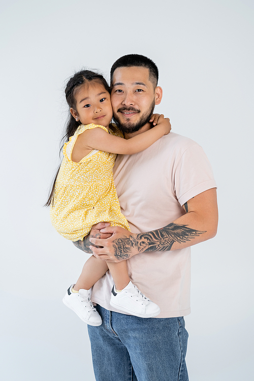 happy and tattooed asian father holding smiling preschooler daughter isolated on grey
