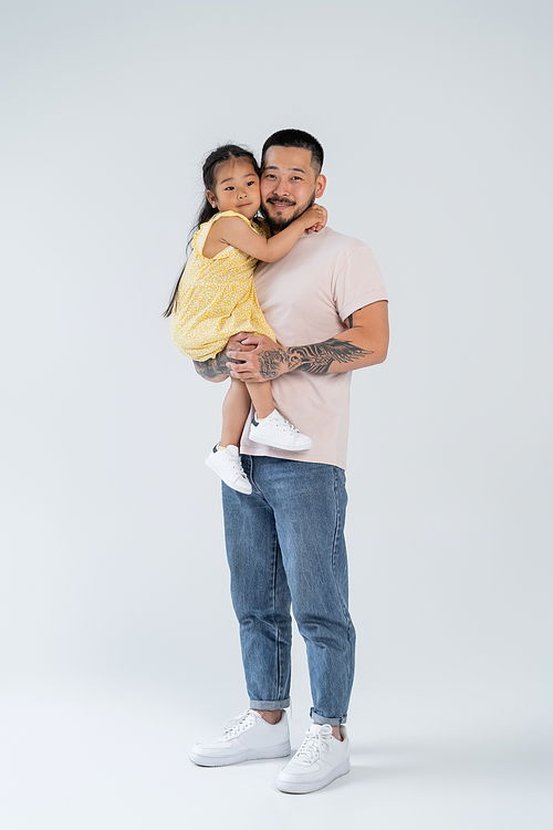 full length of happy and tattooed asian father holding smiling preschooler daughter on grey
