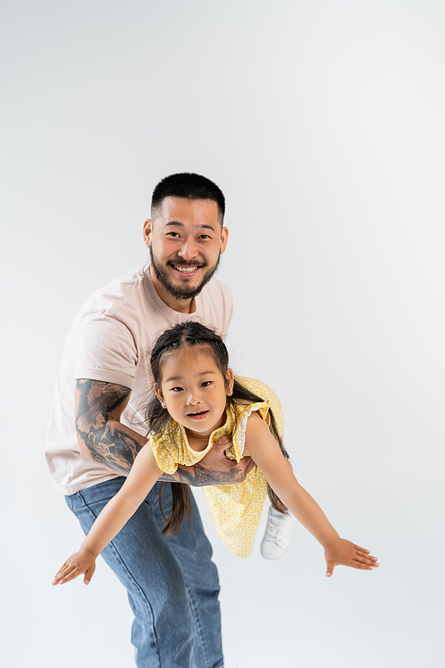 cheerful asian father holding in arms joyful preschooler daughter in yellow dress isolated on grey