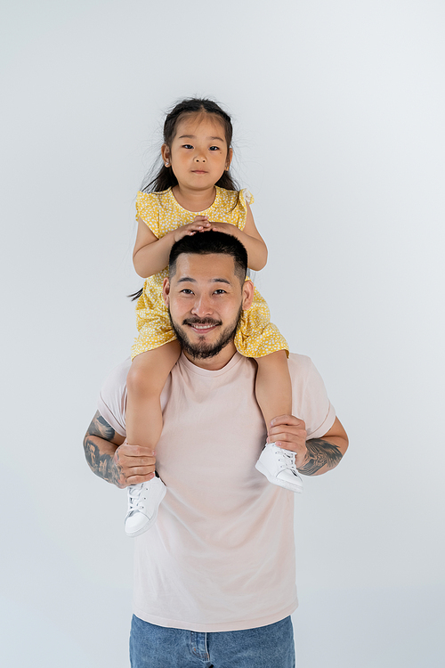 happy asian child sitting on shoulders of tattooed father isolated on grey