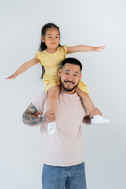 happy asian child with outstretched hands sitting on shoulders of tattooed father isolated on grey