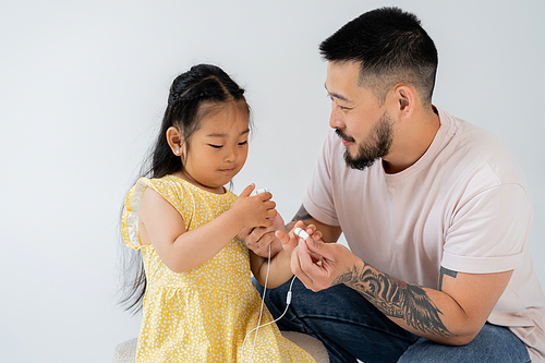 tattooed asian man holding wired earphones near preschooler daughter isolated on grey