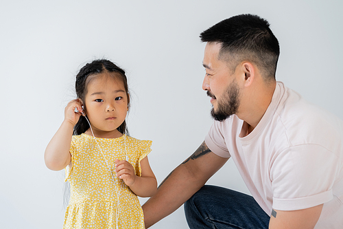 tattooed asian man looking at preschooler daughter wearing wired earphones isolated on grey
