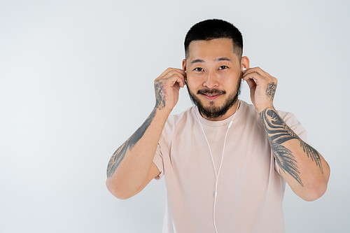 tattooed asian man in t-shirt smiling while wearing wired earphones and  isolated on grey