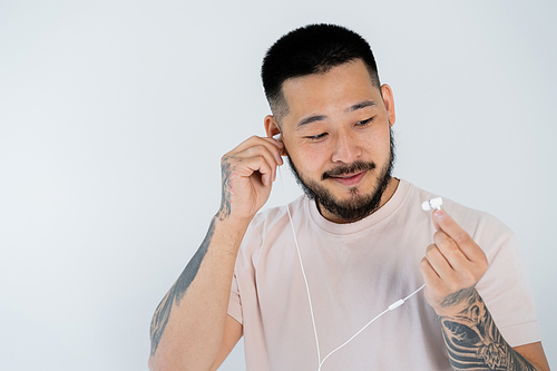 tattooed asian man in t-shirt smiling and wearing wired earphones isolated on grey