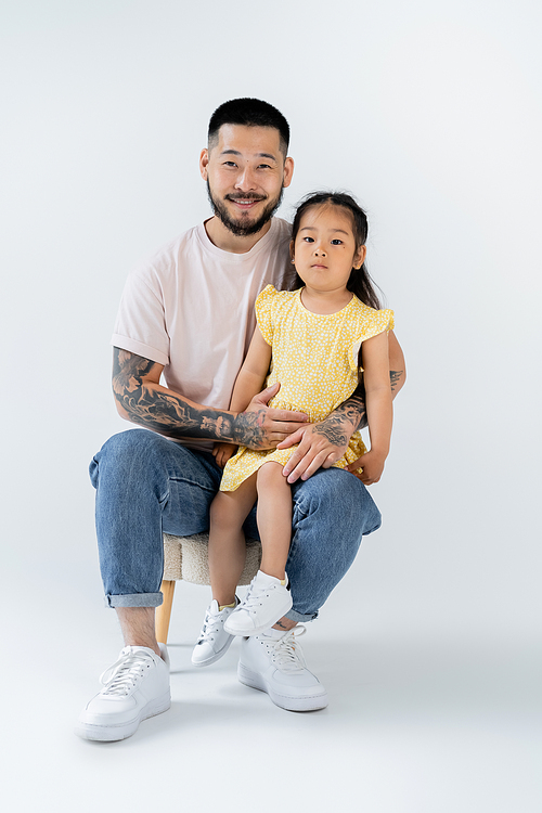 full length of happy asian kid sitting on knee of father in jeans on grey