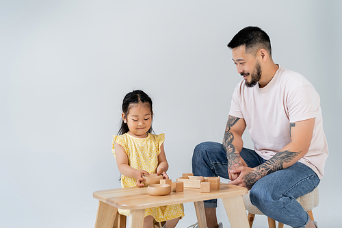 happy asian man looking at brunette daughter playing with wooden toys isolated on grey