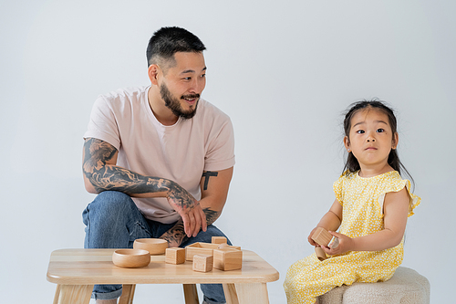 cheerful and tattooed asian man looking at brunette daughter playing with wooden toys isolated on grey