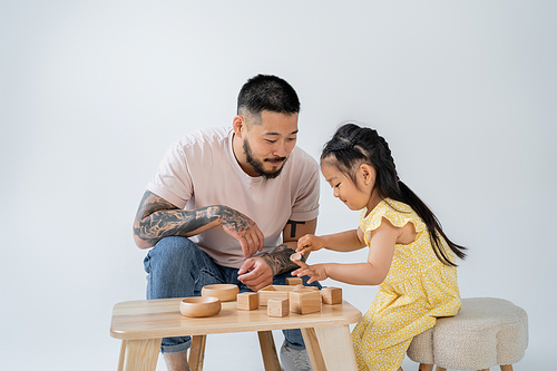 tattooed asian father playing wooden toys with brunette daughter isolated on grey