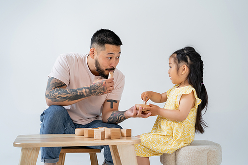 tattooed asian father playing wooden toys with preschooler daughter isolated on grey