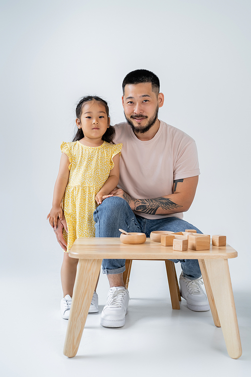 cheerful and tattooed father and asian preschooler daughter near table with wooden toys on grey
