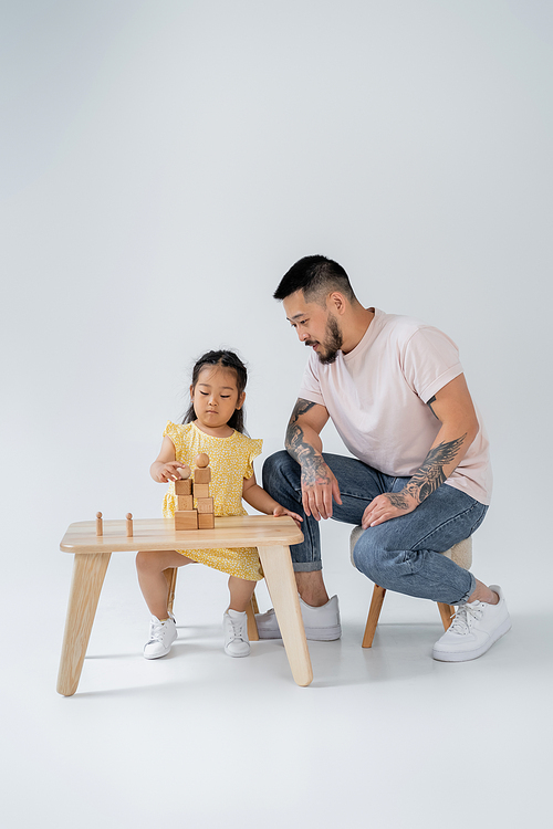 tattooed asian man looking at brunette daughter playing with wooden toys on grey