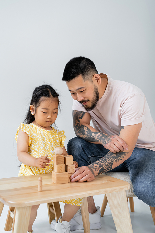 tattooed asian father playing wooden cubes with preschooler daughter isolated on grey