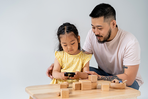 tattooed asian man looking at brunette daughter using smartphone near wooden toys isolated on grey