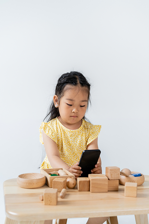 asian girl using smartphone near wooden toys on table isolated on grey
