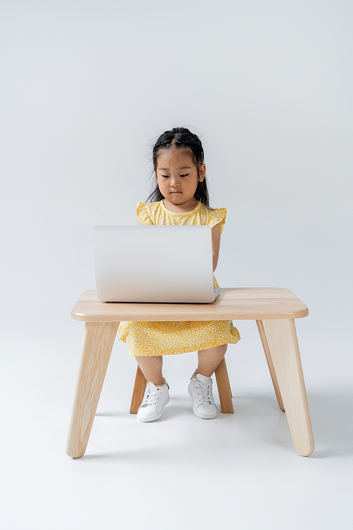 full length of asian girl in yellow dress using laptop while sitting at wooden table on grey