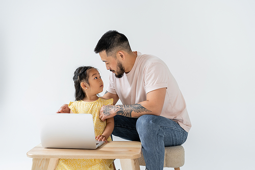 tattooed father looking at surprised asian daughter near laptop on wooden table isolated on grey