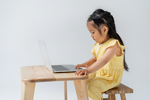 side view of asian preschooler girl in yellow dress using laptop isolated on grey