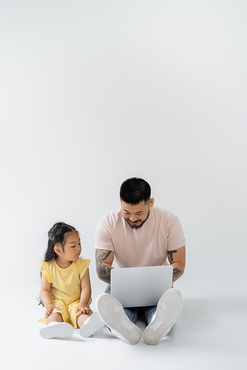 happy asian father working remotely on laptop near daughter on grey