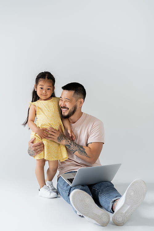 cheerful asian father sitting with laptop near daughter on grey