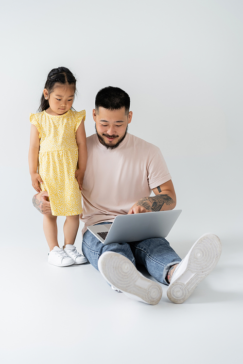 cheerful asian father using laptop near daughter in yellow dress on grey