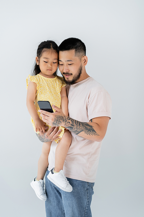 asian tattooed man using smartphone and holding daughter in arms isolated on grey