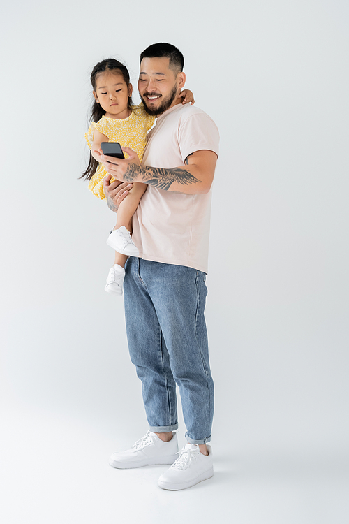 full length of happy tattooed man using smartphone and holding asian daughter in arms on grey