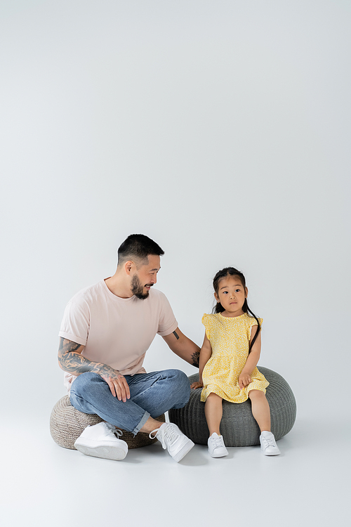 full length of happy tattooed father and asian daughter in yellow dress sitting on puff chairs on grey