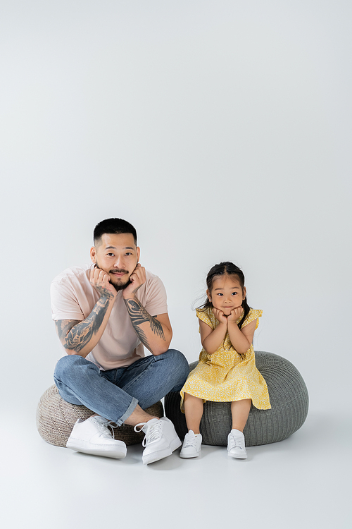 full length of tattooed father and asian daughter in yellow dress sitting on puff chairs on grey