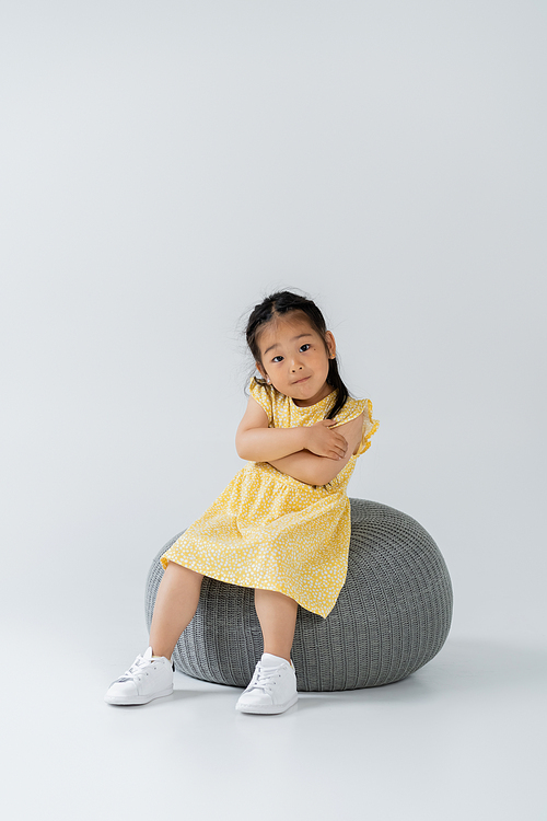 full length of asian kid in yellow dress sitting on puff chair on grey