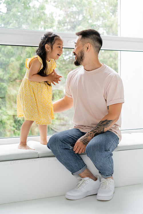 happy tattooed man sitting on windowsill and looking at cheerful asian daughter