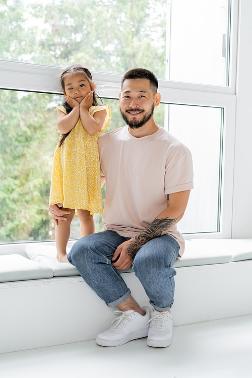 happy tattooed man sitting on windowsill near barefoot and cheerful asian daughter