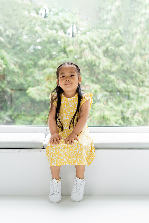 full length of cheerful asian girl in yellow dress sitting on soft pillow on windowsill