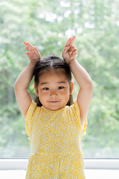 portrait of brunette and preschooler asian girl in yellow dress showing bunny ears near window at home