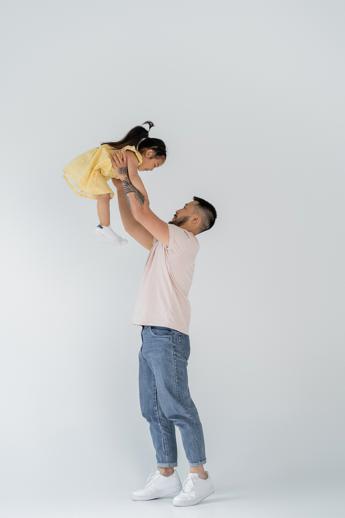 full length of asian man lifting daughter in yellow dress on grey