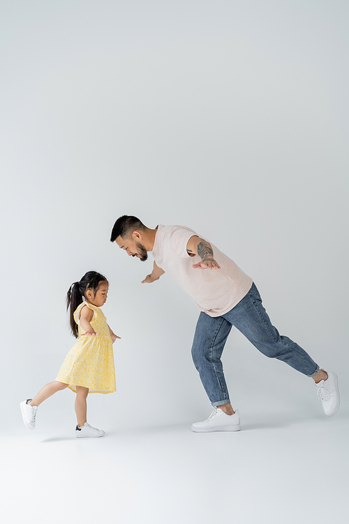 full length of asian father playing with daughter in yellow dress on grey
