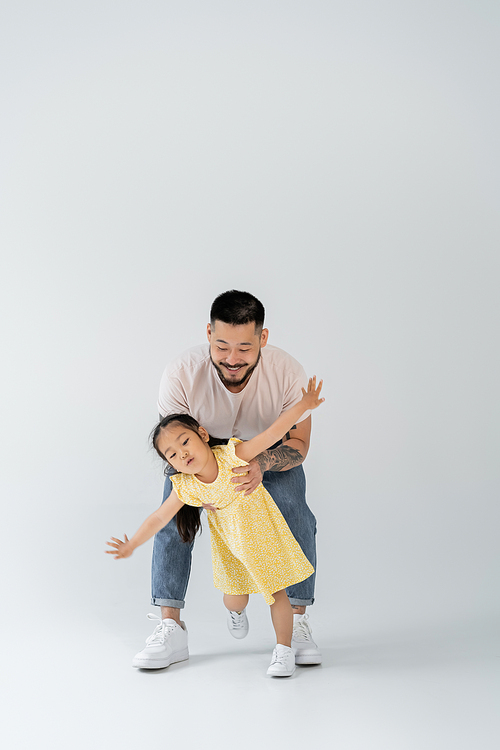 full length of happy asian father playing with daughter in yellow dress standing with outstretched hands on grey