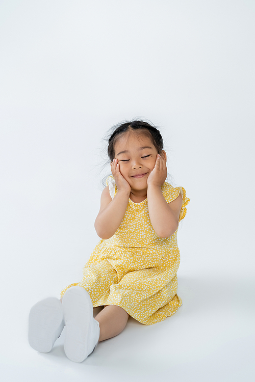 full length of cute asian child in yellow dress smiling with closed eyes while sitting on grey