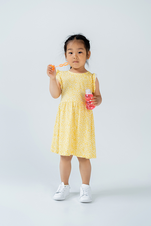 full length of asian girl in yellow dress holding bottle with soap bubbles on grey