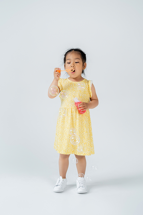 full length of asian preschooler kid in yellow dress holding bottle with soap bubbles on grey