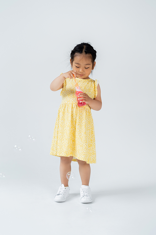 full length of asian preschooler child in yellow dress holding bottle with soap bubbles on grey